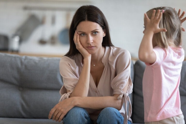 Unhappy mother having problem with noisy naughty little daughter screaming, demanding attention, child tantrum manipulation concept, tired thoughtful mum holding head, sitting on couch at home