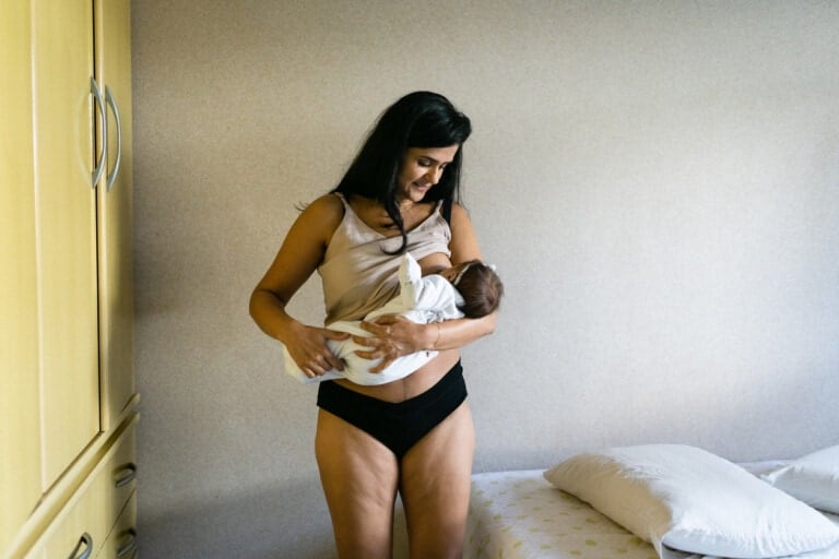 A woman stands in a room holding a baby in her arms, navigating the after birth symptoms. She is wearing a light-colored top and black underwear. The room has beige walls, a bed with a white pillow, and a yellow wardrobe on the left side of the image. The woman is looking down at the baby.