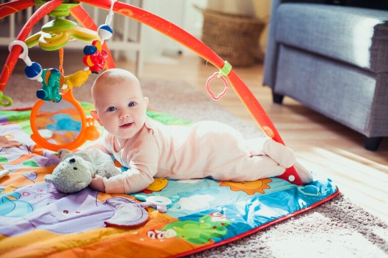 3 months old baby girl lying on colorful play mat on the floor. Activity carpet for kids. Early development at home.