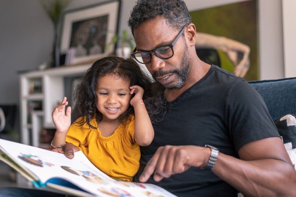 A loving father of African descent sits on the couch at home and reads a storybook to his preschool age daughter. The child is sitting on her father's lap and is smiling while looking at the book.
