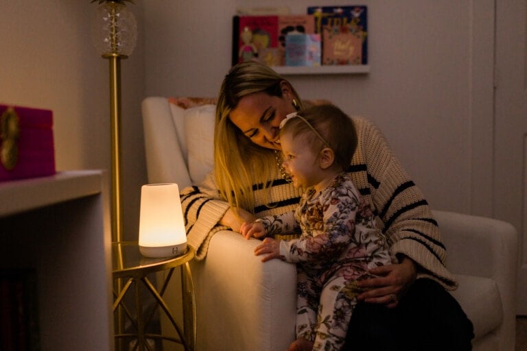 Mother sitting in her daughter's nursery with her daughter on her lap as they look at the glow of the Hatch Rest with the lights off in the room.