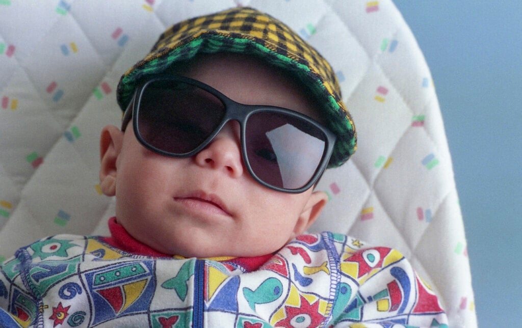 One cute Latino baby boy lying on tabletop baby bouncer wearing a checkered cap and sunglasses.