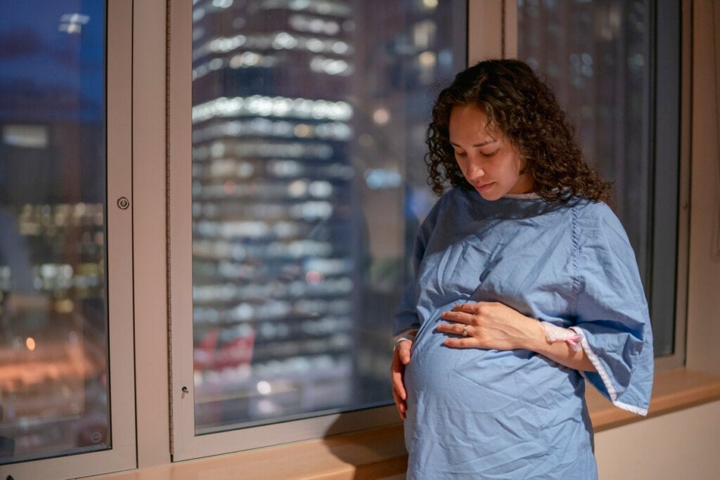 An ethnic soon-to-be mother is wearing a hospital gown and staring down at her pregnant belly while leaning against a window during the twilight night hours.