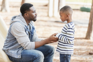 An African-American man in his 30s with a serious expression on his face, talking to his 7 year old son on a playground. They are face to face, and he is holding his hands.