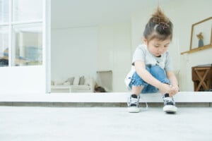 Young child sits on a doorstep, looking down while tying shoelaces, with a bright, minimal living room visible in the background.