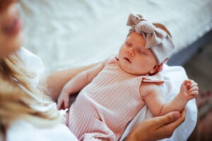 Young mother holding her sleeping baby girl in her lap. Baby is wearing a onesie and a bow headband.