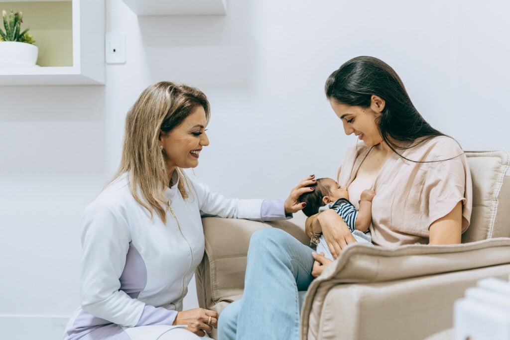 Nurse doula accompanying mother and baby while she breastfeeds sitting in a chair.
