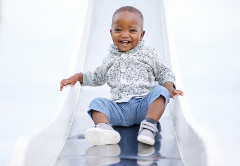 Baby boy smiling big sitting on the end of a slide.