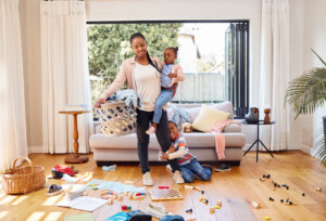 Mom walking through a messy living room holding her daughter and her son clinging to her leg. Mom looks overwhelmed.