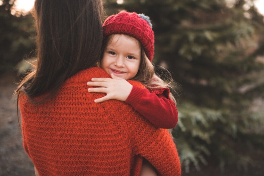 Cute smiling child girl 3-4 year old on mother hands wear knit hat and sweater outdoor. Look at camera. Christmas holiday season.