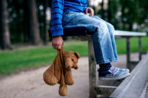A child, wearing a blue striped shirt and ripped jeans, sits on a bench holding a brown teddy bear by its arm. The child's face is not visible. Trees and a grassy area are in the background, evoking thoughts of what to teach kids to do when they are lost.