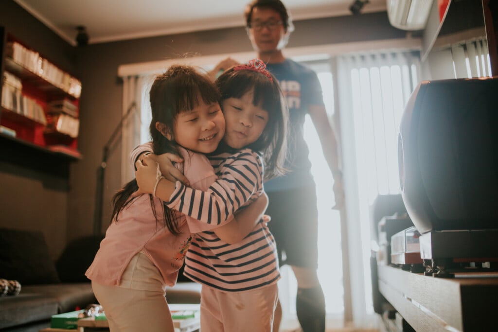 Two sisters standing in the living room giving each other a hug while their dad is standing in the background.