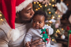 A man wearing a Santa hat holds and kisses a baby dressed in a gray sweater with a Christmas tree design, capturing photos of baby's first Christmas. They are in front of a decorated Christmas tree with lights.