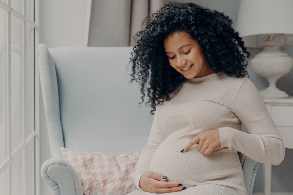 Women and pregnancy concept. Beautiful happy pregnant woman in white dress with curly hair sitting on comfortable armchair at home near window and pointing at her future baby in her belly.