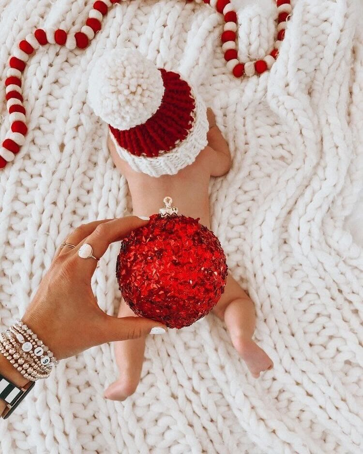 Woman's hand holding a christmas ornament covering a baby's bottom. Baby is laying on a blanket wearing a santa's hat.