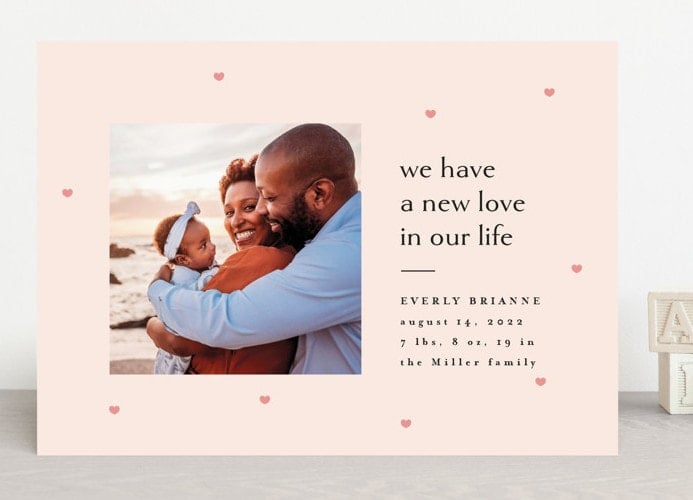 Pink rectangle card with a photo of parents holding a baby and text announcing the baby on the right side of the card. 