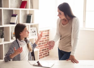 A young girl with headphones around her neck and an older woman, both with expressive gestures, appear to be having a serious conversation in a room with shelves and a window. A tablet lies on the table between them, hinting at underlying power struggles.