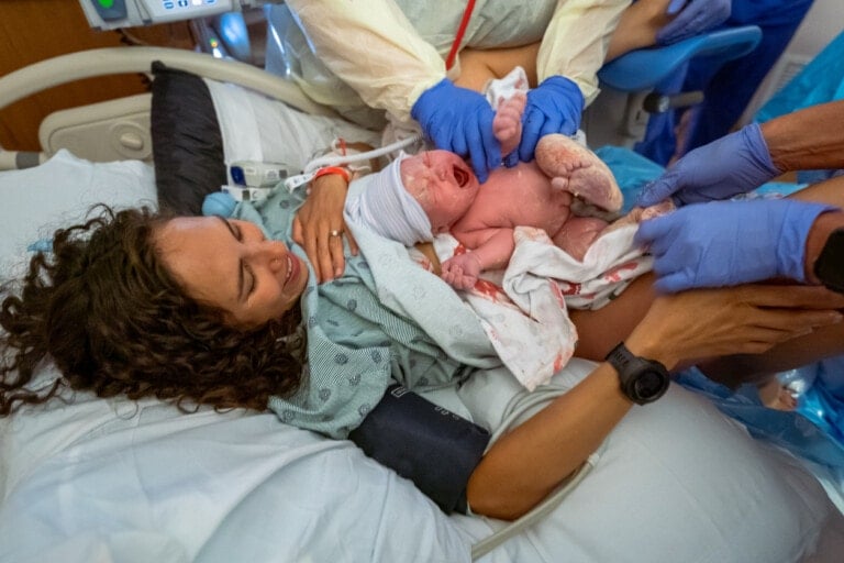 A multiracial woman smiles with gratitude as her doctor and nurse place her healthy newborn baby on her chest for skin to skin bonding directly after vaginal delivery birth.