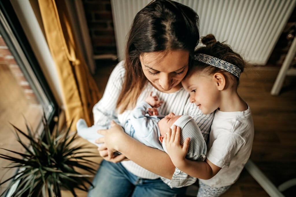Overhead shot of a mother holding her new baby while she is sitting down and her daughter standing next to her resting her head on her shoulder as she looks down at the baby smiling.