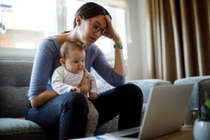 Tired young mother working from home holding her baby in her lap while looking at her computer.