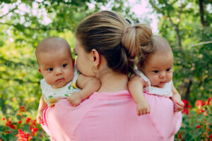 Young Caucasian mother holding identical twin girls in the hand in the garden with flowers