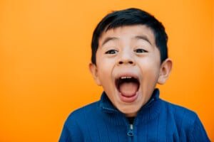 A child with short dark hair and a blue sweater is pictured against an orange background. His mouth is wide open, and his eyes are wide, giving an impression of excitement or surprise—perhaps he just heard some funny jokes for kids!