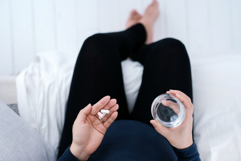 Personal perspective of Asian pregnant woman sitting on bed side at home, taking medicines in hand with a glass of water. Essential vitamins and supplements are vital for the healthy growth and development of the unborn baby