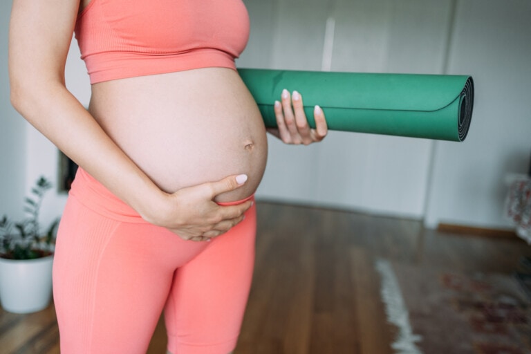 Cropped shot of young unrecognizable pregnant woman practicing yoga at home. Pregnant woman holding exercise mat in one hand and touching her belly with other hand.