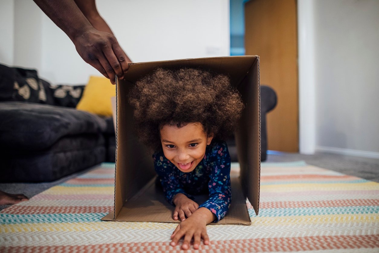 Mixed race girl crawling through a cardboard box in the sitting room at home, her unrecognizable father is holding the box up for her.