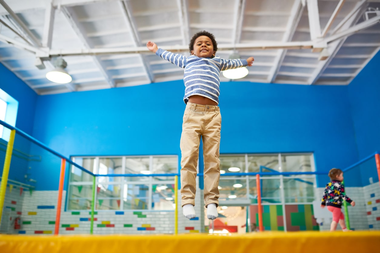 Full length low angle portrait of happy African-American boy jumping on trampoline in colorful kids play center