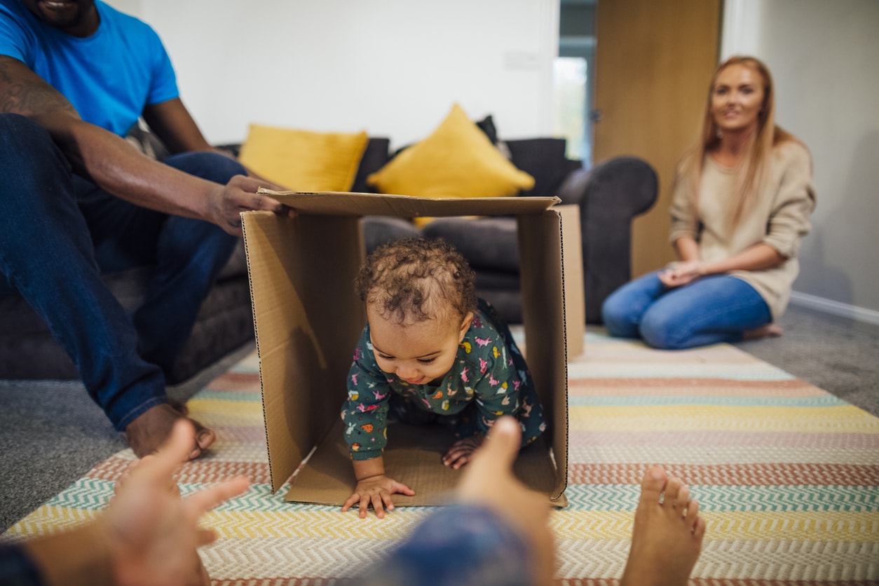Mixed race baby girl crawling through a cardboard box in the sitting room at home, her father is holding the box up for her, her sister is holding her hands out for her to come to her and her mother is in the background.