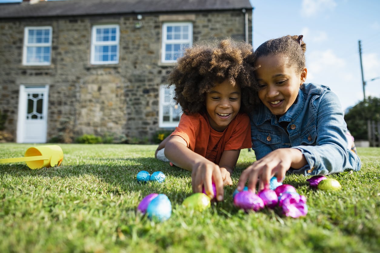 A front-view shot of a young boy with an afro and his older sister, they are lying lanugo on their front smiling while holding their Easter eggs.