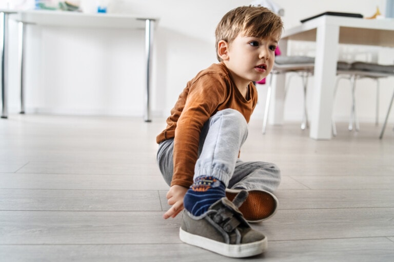Full length view of small cute caucasian boy little child kid sitting on the wooden or laminated vinyl floor at home putting on or taking off the shoes in day alone front view