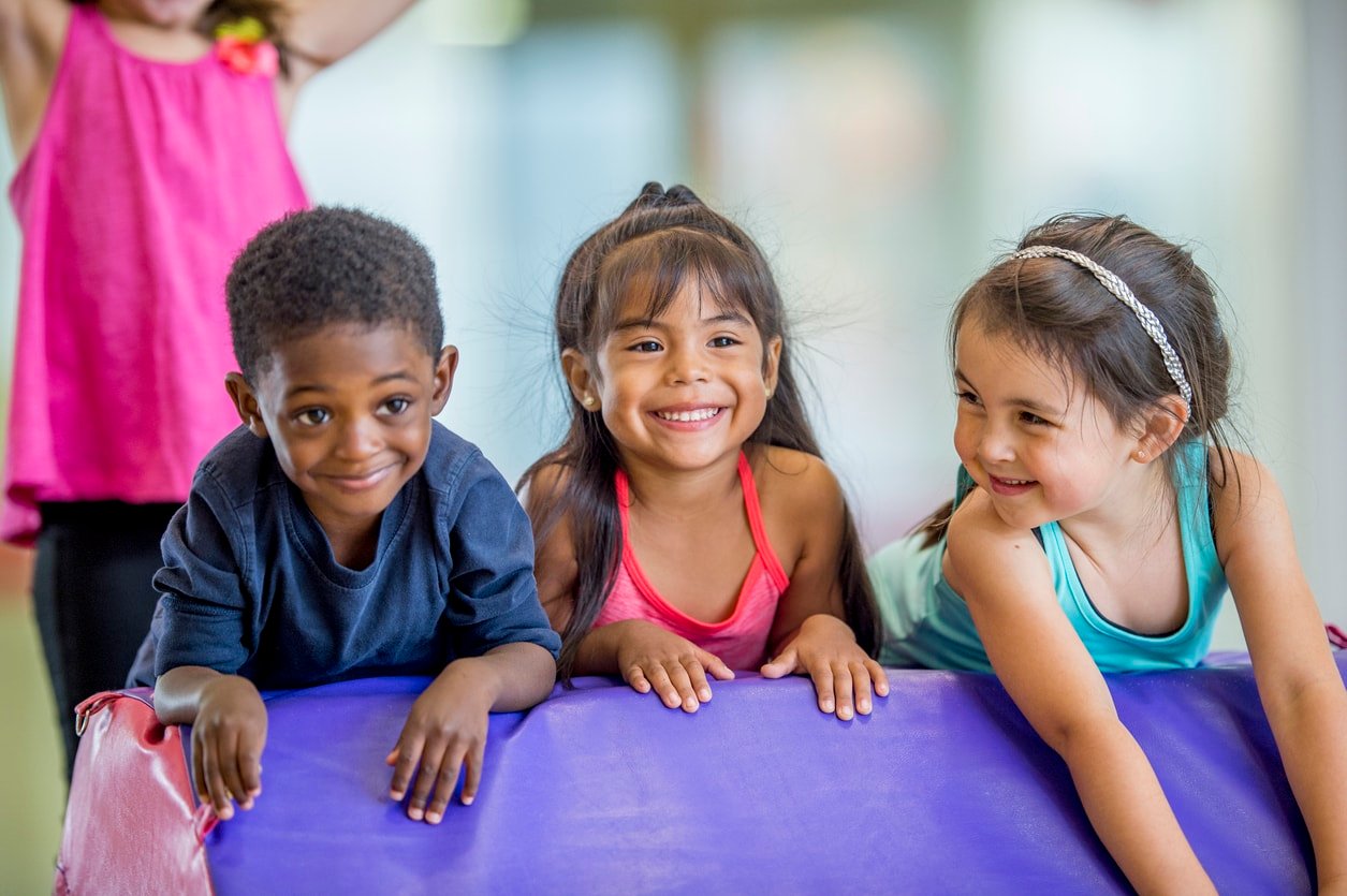 Three preschool kids are indoors in their school. They are having fun during gym class. They are leaning on a gymnastics mat and smiling together.