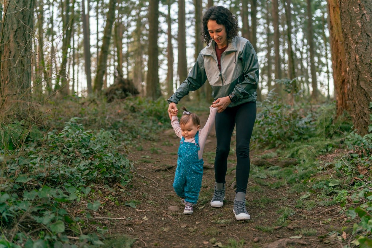 A gorgeous mixed race Pacific Islander mother is holding her one year old baby daughter's hands and helping the young child learn to walk. The family is on an adventurous walk in the forest at sunset.