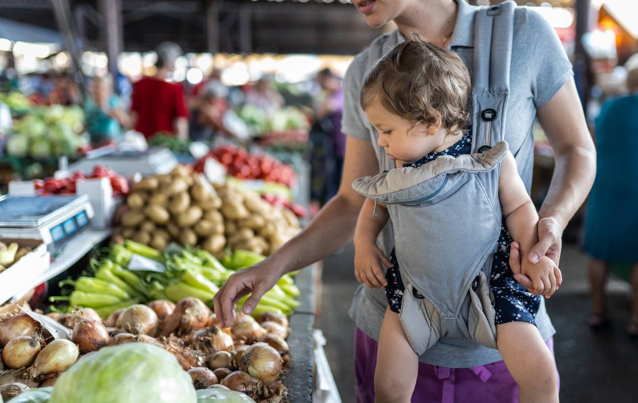 Young mother carrying her cute girl in baby carrier and looking vegetables on farmer's market