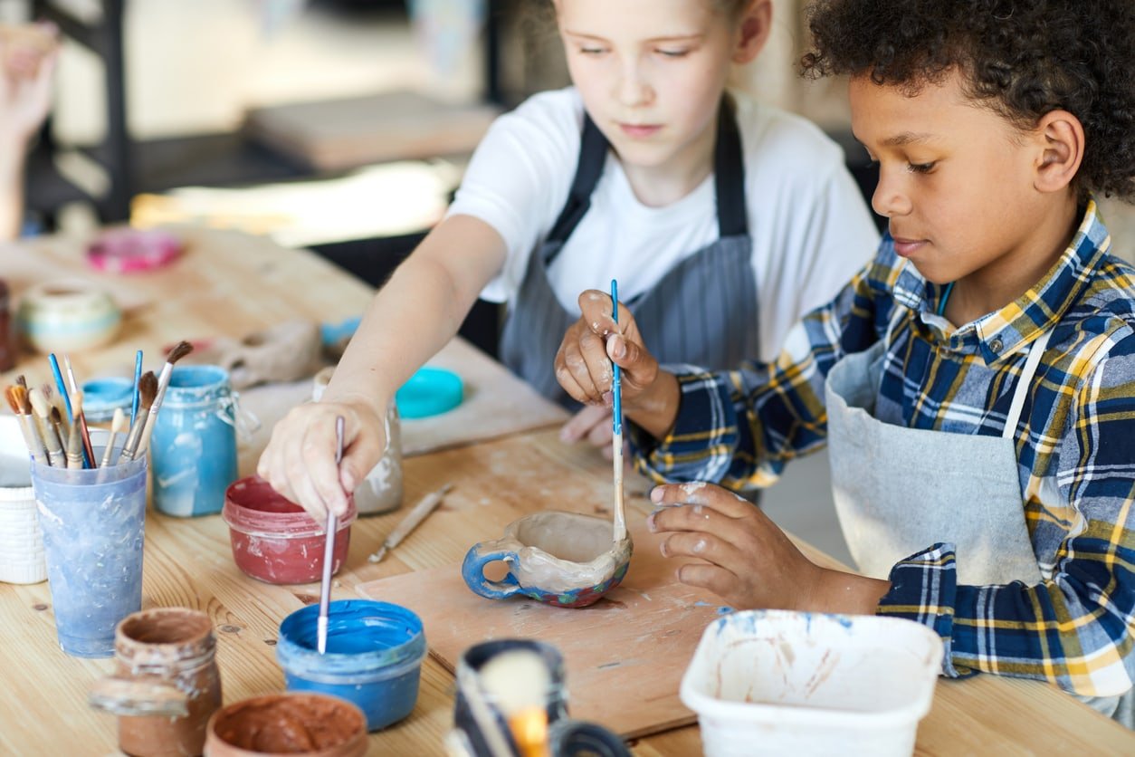 Two youthful classmates sitting by table and painting self-made clay items at lesson