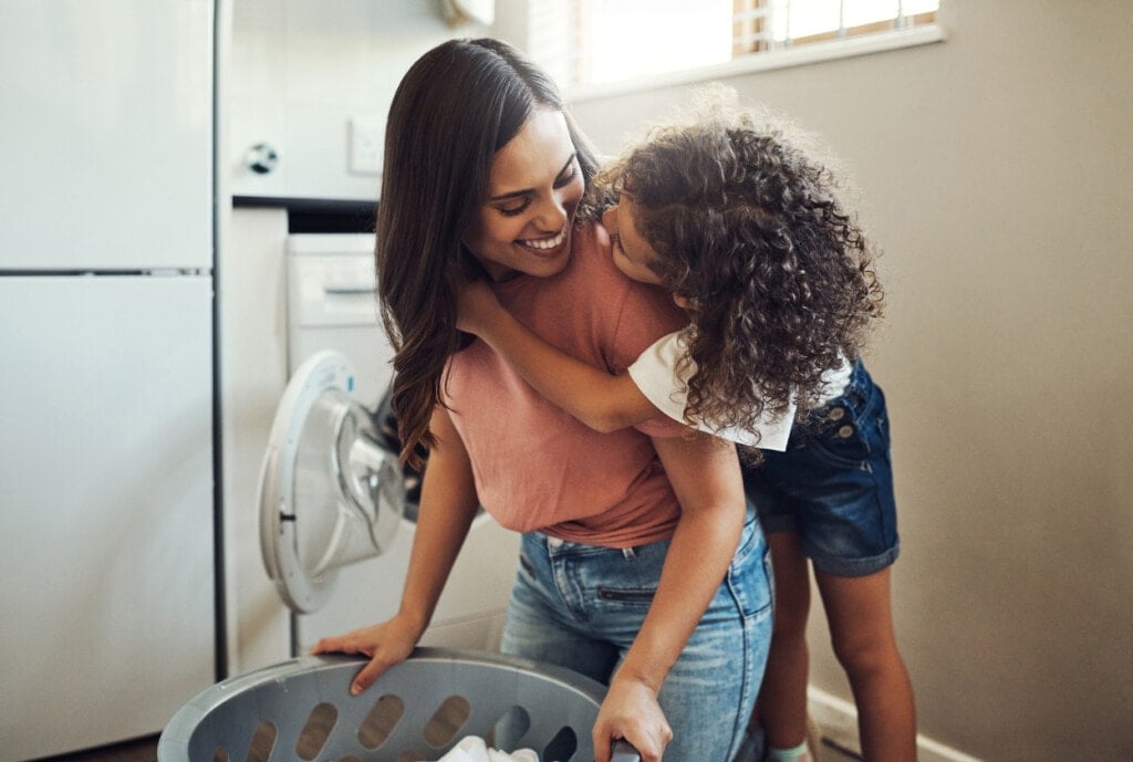 Mom and young daugther are in the laundry room. Mom is kneeling down holding on to the laundry basket and the daughter has her arms around her mother's neck standing behind her.