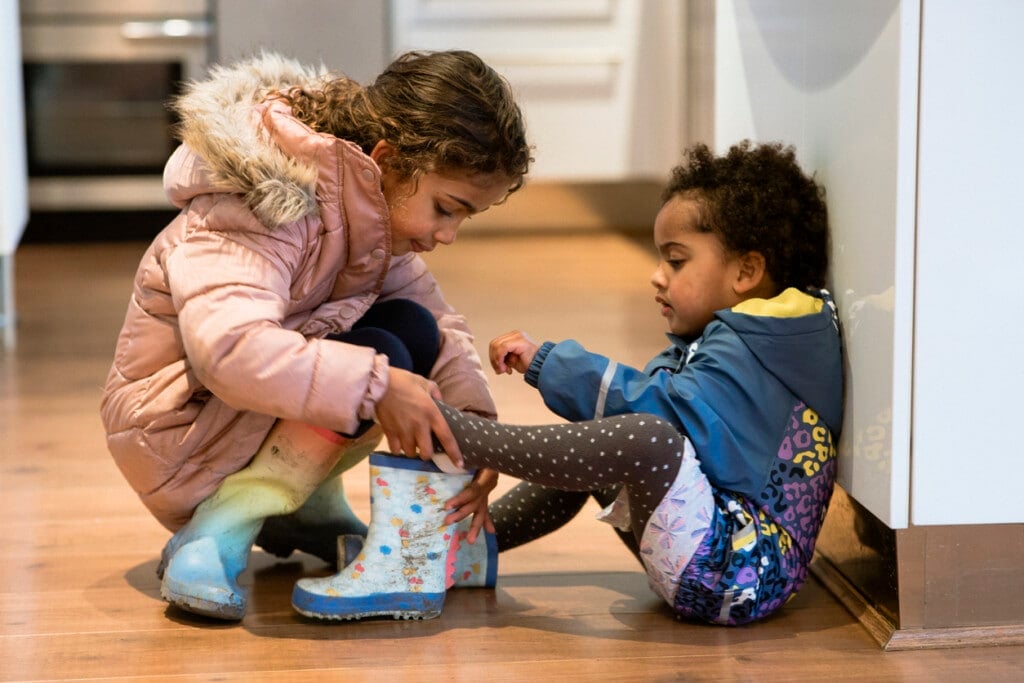 Mixed race sisters, dressed in their warm coats, getting ready to go outside and play. The older sibling is helping the younger sister put on her wellington boots.