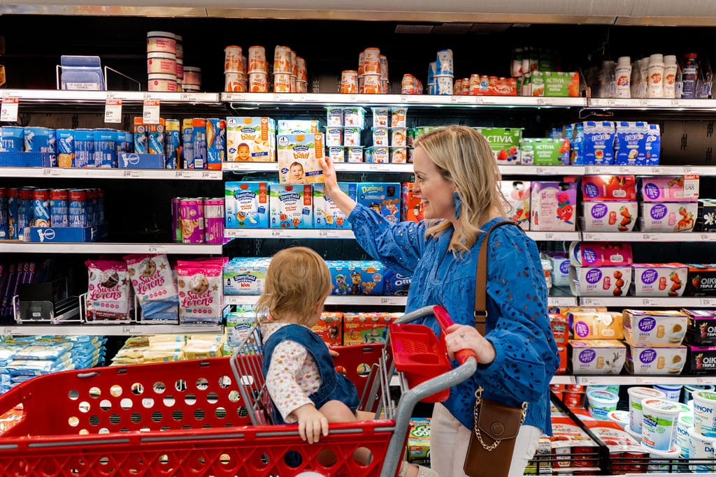 Mom shopping with daughter at Target. Daughter is in the cart and they are looking at yogurt. The mom is reaching for Stonyfield Yobaby yogurt.