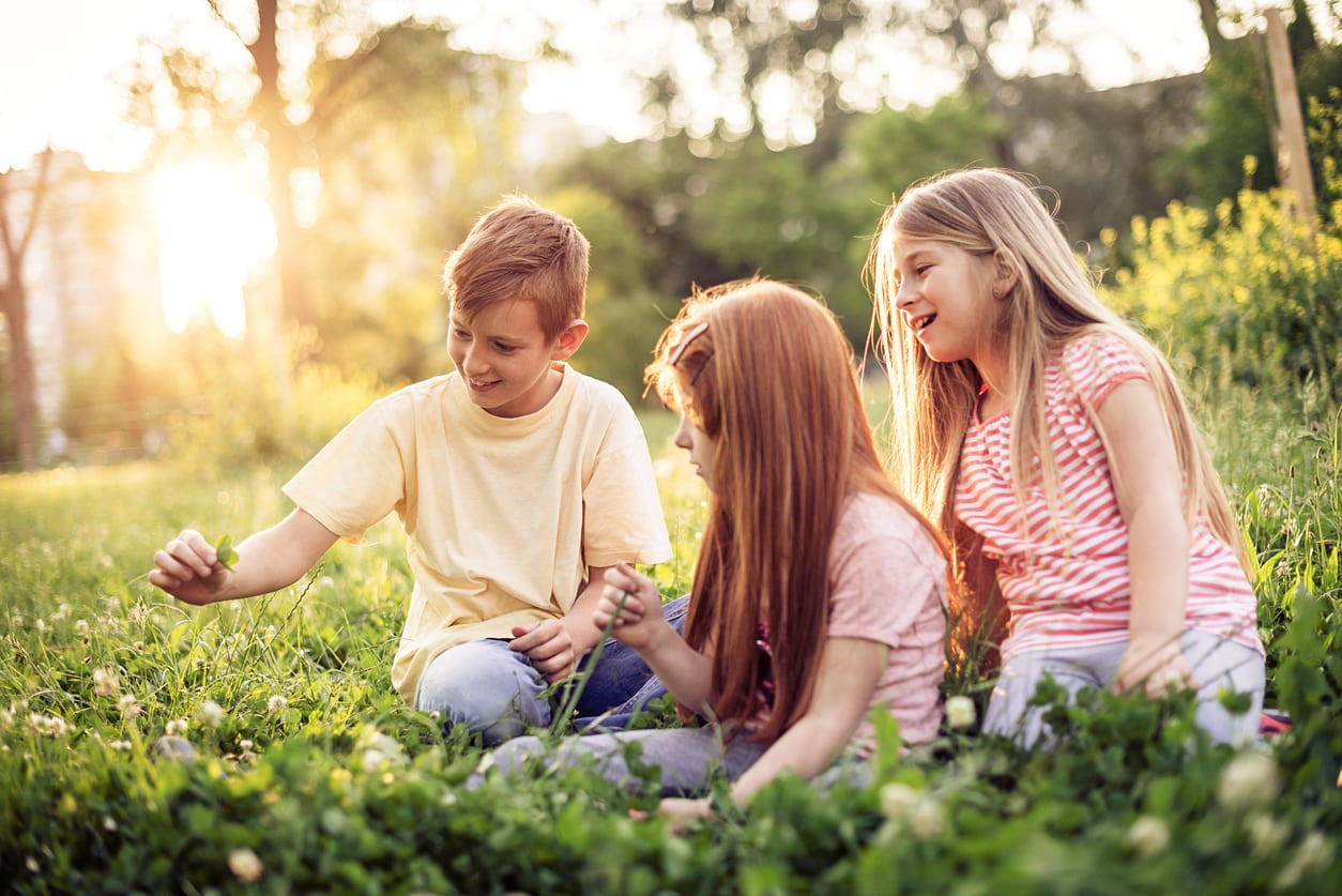 Three children sitting outside in a field of grass looking for four leafed clovers.