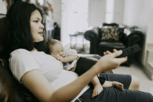 A close-up of a mother holding a newborn baby son at home. She's holding a TV remote control probably looking at the TV as her baby lays on her best and belly asleep.
