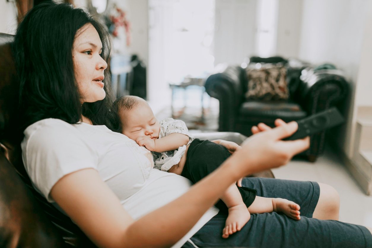 A close-up of a mother holding a newborn baby son at home. She's holding a TV remote control probably looking at the TV as her baby lays on her best and belly asleep.