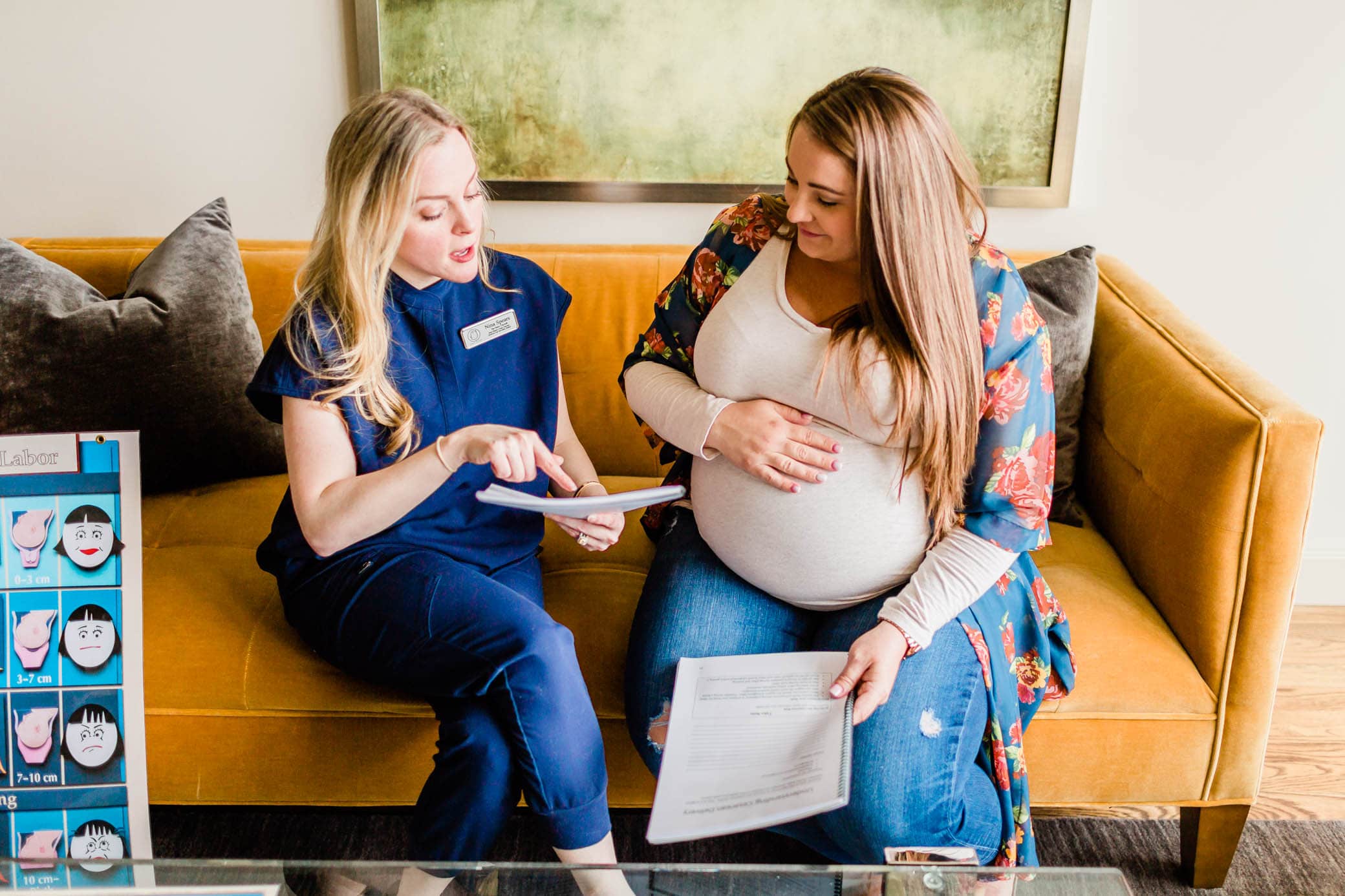 A doula sitting down with her client, a pregnancy woman, on the couch and they are looking over some printed papers and talking.