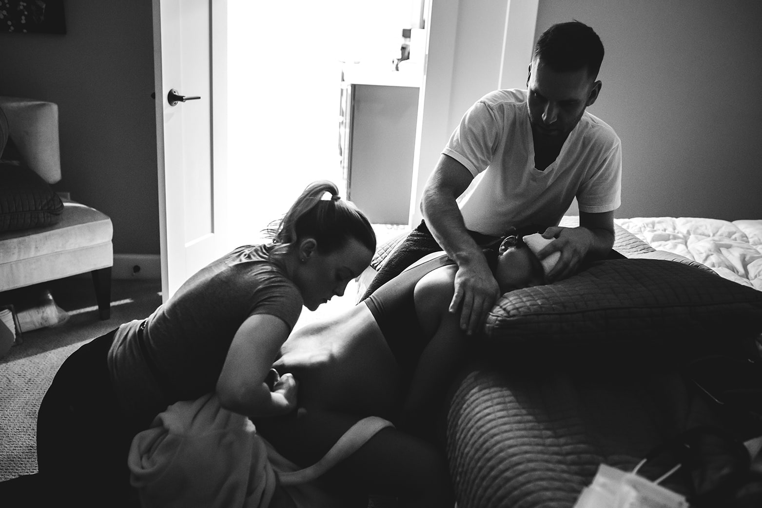 Laboring woman sitting on a birthing ball in her bedroom. Her husband is by her head supporting her and she has a doula behind her doula counter pressure. It's a black and white photo.