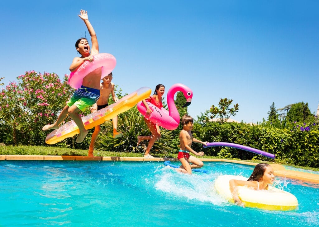 Group of age-diverse boys and girls, happy friends with swim floats jumping into swimming pool on vacation.