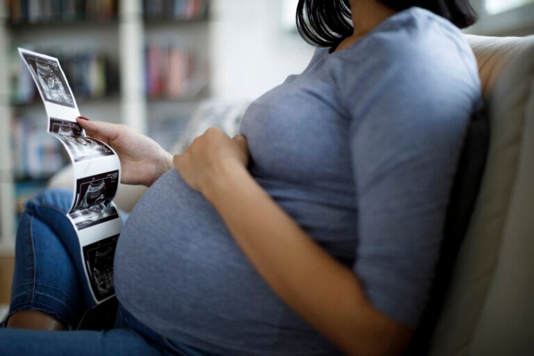 Pregnant woman looking at ultrasound scan of baby while sitting on a couch.