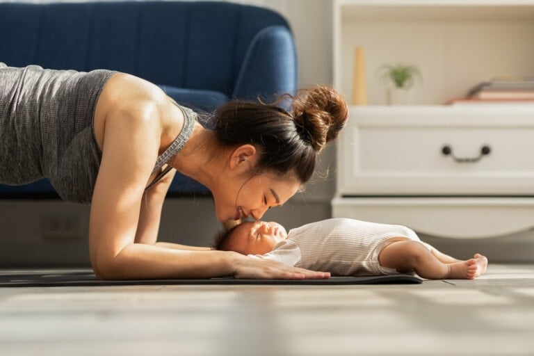 Side view of Asian woman mom doing plank exercise and kissing her baby who is laying on the floor at home. Happy healthy mother yoga plank with newborn baby boy sleep and lying on yoga mat.