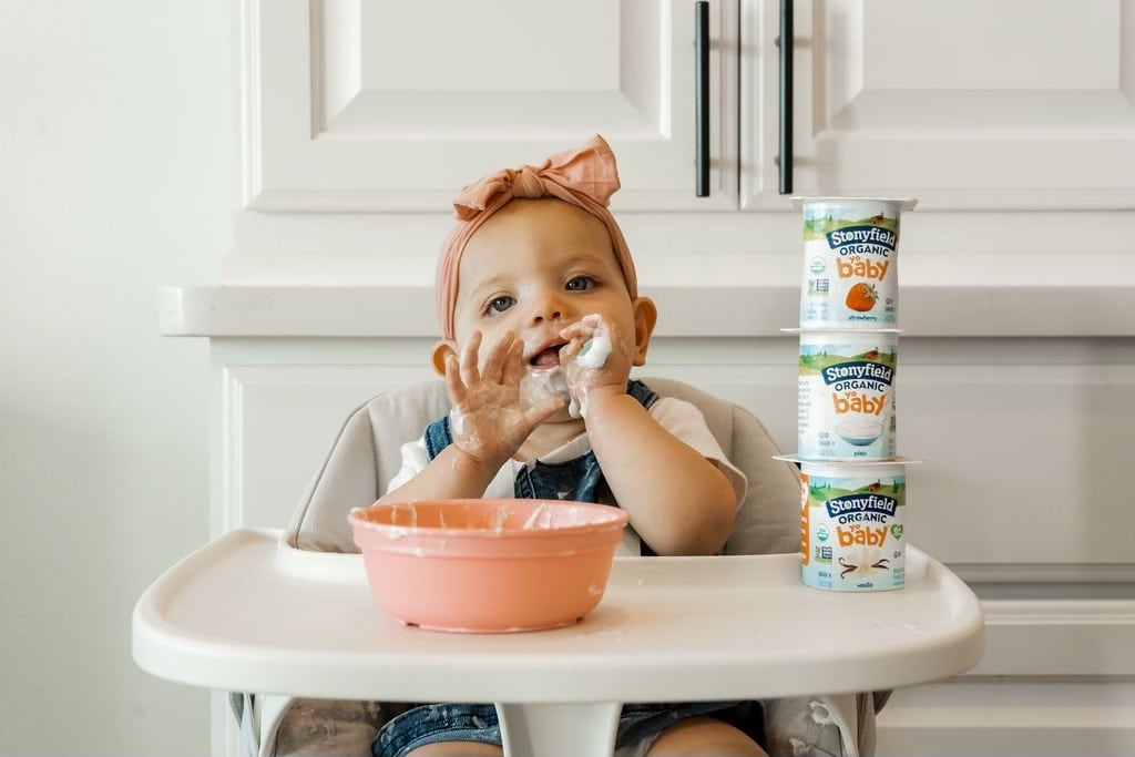 Baby sitting in highchair eating yogurt out of a bowl. 3 Stonyfield Baby Yogurt cups stacked on top of each other next to baby.