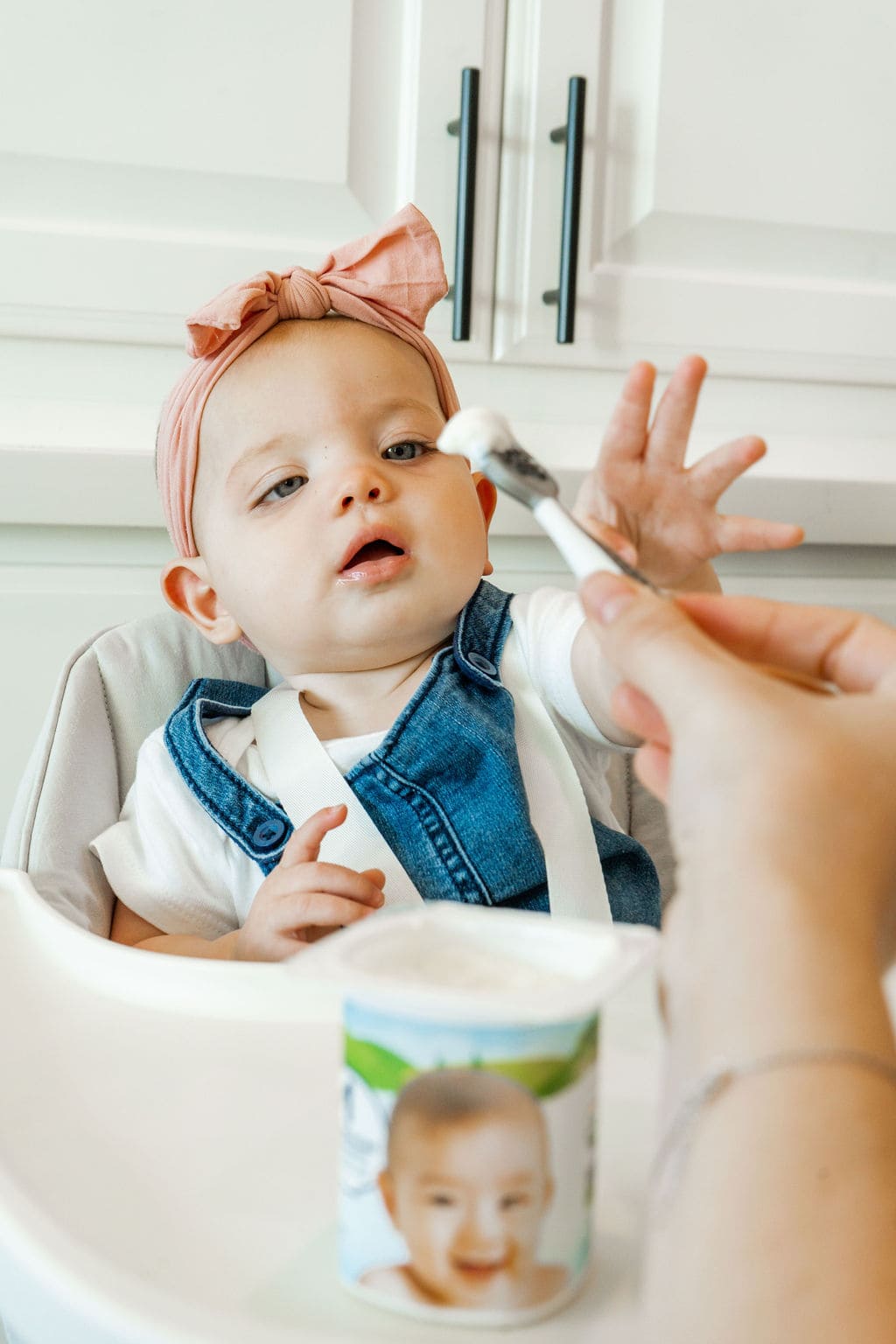 A spoon with yogurt on it and a hand giving it to the baby girl in her high chair.